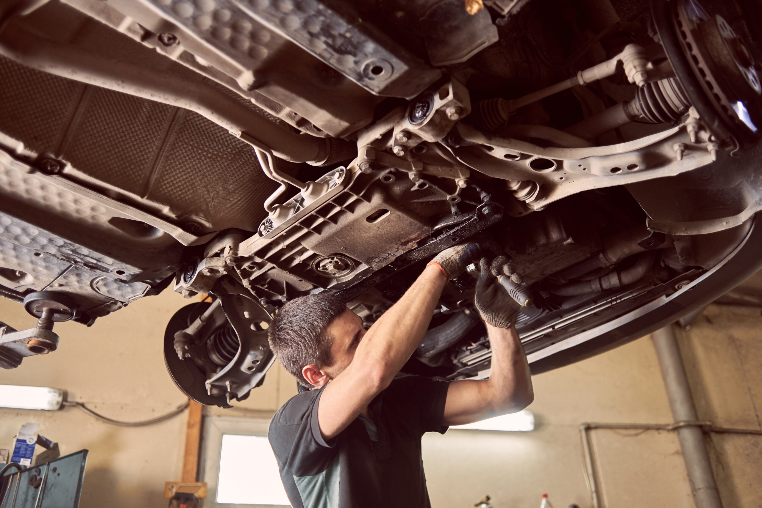 Repairman fixing car in garage. Experienced specialist car mechanic standing under lifted car during repair and maintenance process in repair station. Workshop for automobile checking up and repairing