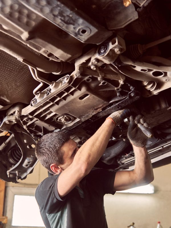 Repairman fixing car in garage. Experienced specialist car mechanic standing under lifted car during repair and maintenance process in repair station. Workshop for automobile checking up and repairing